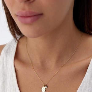 May include: A gold-toned chain necklace featuring two small pendants: one oval-shaped charm with an engraved floral design and a second smaller circular charm set with a single dark blue stone. The necklace rests against a white textured top.