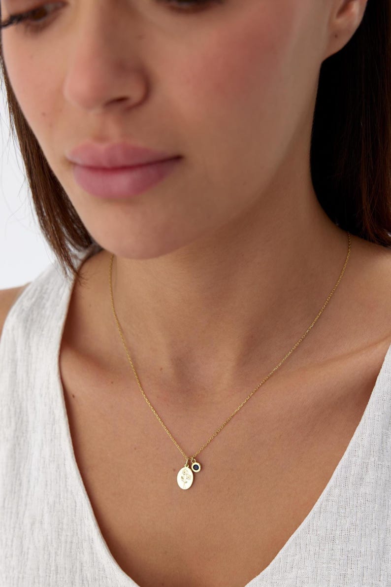 May include: A gold-toned chain necklace featuring two small pendants: one oval-shaped charm with an engraved floral design and a second smaller circular charm set with a single dark blue stone. The necklace rests against a white textured top.