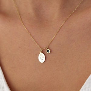 May include: A gold-toned necklace featuring a delicate chain with two pendants: an oval-shaped charm engraved with a fine-line floral illustration and a small, circular charm set with a dark, round stone.