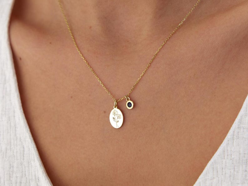 May include: A gold-toned necklace featuring a delicate chain with two pendants: an oval-shaped charm engraved with a fine-line floral illustration and a small, circular charm set with a dark, round stone.