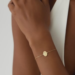 Puede incluir: Una pulsera de tono dorado con una cadena delicada y un dije central de forma ovalada. El dije presenta un grabado de una ilustraci&oacute;n bot&aacute;nica de l&iacute;neas finas de una flor con tallo y hojas, centrado en la superficie de metal pulido.