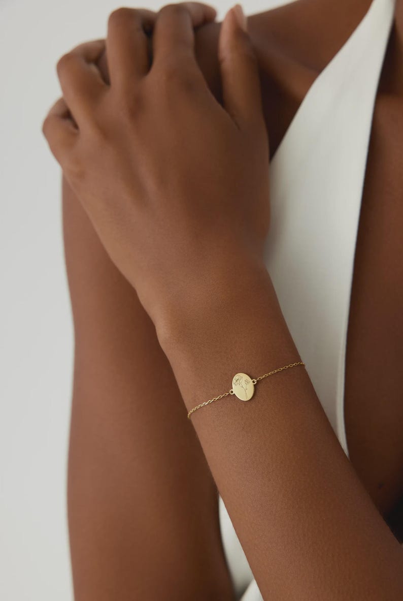 Puede incluir: Una pulsera de tono dorado con una cadena delicada y un dije central de forma ovalada. El dije presenta un grabado de una ilustraci&oacute;n bot&aacute;nica de l&iacute;neas finas de una flor con tallo y hojas, centrado en la superficie de metal pulido.