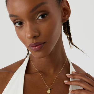 May include: A gold-toned chain necklace featuring two small pendants: one oval-shaped charm with an engraved floral design and a second smaller circular charm with a dark, round center stone. The necklace rests against a white sleeveless garment.