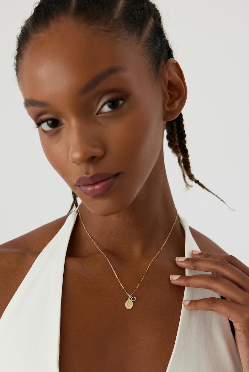 May include: A gold-toned chain necklace featuring two small pendants: one oval-shaped charm with an engraved floral design and a second smaller circular charm with a dark, round center stone. The necklace rests against a white sleeveless garment.