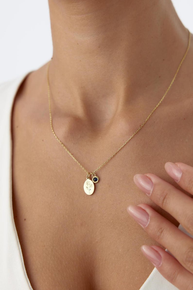 May include: A gold chain necklace featuring a small oval pendant engraved with a floral design and a separate circular charm set with a single dark blue gemstone. The delicate metal chain rests against the neck, showcasing the two hanging charms.
