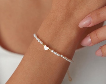 May include: A delicate bracelet featuring a single row of small, white, irregularly shaped freshwater pearls. A tiny, polished gold-tone heart charm is positioned at the center of the pearl strand, flanked by two small gold-tone beads.