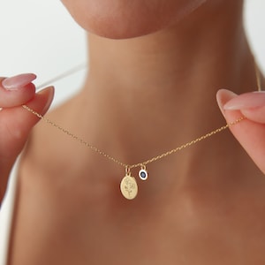 May include: A gold chain necklace featuring two small pendants: an oval-shaped charm engraved with a delicate line drawing of a flower and a circular charm set with a single, small, dark blue gemstone. The necklace has a thin, polished gold link chain.