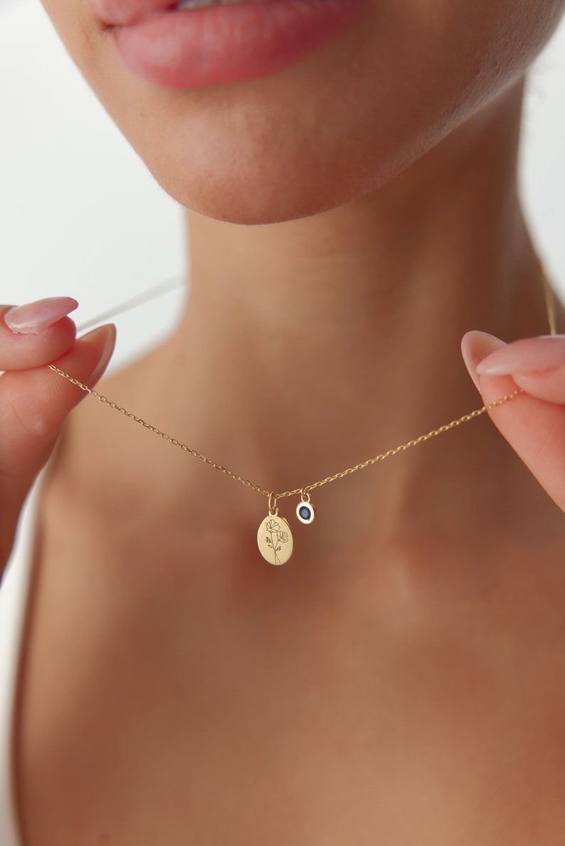 May include: A gold chain necklace featuring two small pendants: an oval-shaped charm engraved with a delicate line drawing of a flower and a circular charm set with a single, small, dark blue gemstone. The necklace has a thin, polished gold link chain.