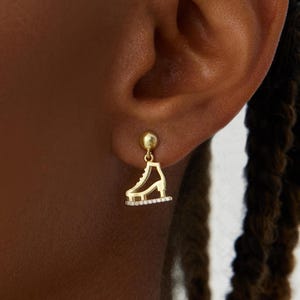 以下が含まれることがあります： クリアなクリスタルアウトラインが付いたゴールドトーンのアイススケートイヤリング。イヤリングは小さなゴールドボールからぶら下がっています。