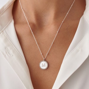 May include: A silver necklace with a round pendant featuring the letter "M" in a raised design. The pendant has a textured edge.