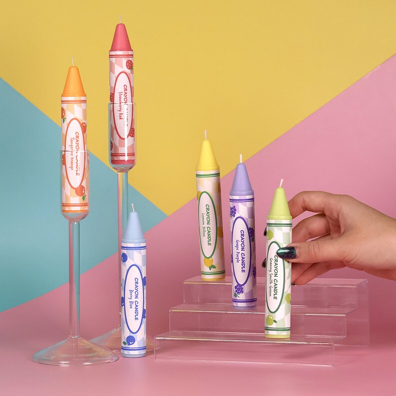 May include: Six colorful crayon candles, each with a different fruit scent. The candles are arranged on a tiered acrylic display stand. The scents are: Strawberry Rad, Tangerine Orange, Granny Smith Green, Grape Purple, Lemon Yellow, and Berry Blue.