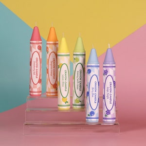May include: Six colorful crayon candles arranged on a tiered acrylic display. The candles are labeled with the scents: Strawberry Red, Tangerine Orange, Lemon Yellow, Granny Smith Green, Berry Blue, and Grape Purple.