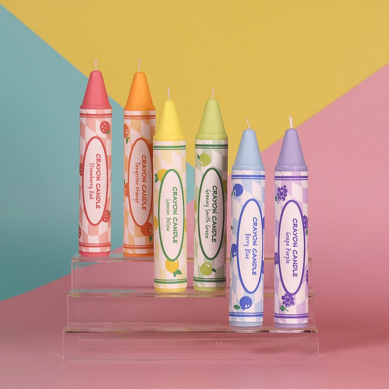 May include: Six colorful crayon candles arranged on a tiered acrylic display. The candles are labeled with the scents: Strawberry Red, Tangerine Orange, Lemon Yellow, Granny Smith Green, Berry Blue, and Grape Purple.