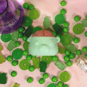 May include: A green and pink gummy bear with a brain on top. The bear has a stitched-up face and a bandage on its eye. The bear is surrounded by green gummy candies.
