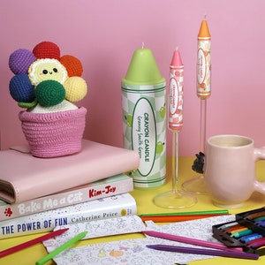 May include: A crocheted flower pot with a rainbow of colors and a cute face. The pot is sitting on a stack of books with the titles "Bake Me a Cat" and "The Power of Fun". There are also crayon candles, coloring pencils, and a mug in the image.