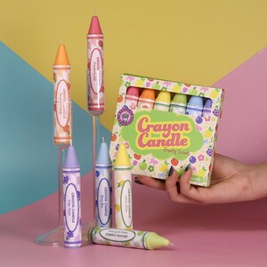 May include: A set of crayon-shaped candles in a box with a colorful design. The candles are labeled with different fruit scents, including strawberry, tangerine, lemon, berry, and grape. The box says "Crayon Candle" and "Fruity Scents".