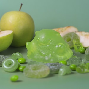 Könnte beinhalten: Ein grünes Gummibärchen in Monsterform mit einem lächelnden Gesicht, umgeben von grünen Gummibärchen, grünen Erbsen und grünen Apfelscheiben.