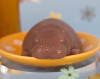 May include: A purple hippopotamus-shaped soap bar on a yellow saucer. The soap is sitting on a glass surface with colourful paper flowers scattered around it.
