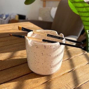 May include: A speckled white ceramic brush holder with two paint brushes inside. The brushes have black handles and light-coloured bristles. The holder has the text "Keep Smiling" on the side.