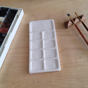 May include: A white ceramic watercolor palette with ten rectangular wells, positioned on a light wood surface. A box of watercolor paints and two paint brushes are also visible. The palette is designed for mixing paints.