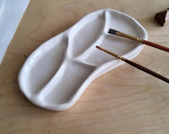 May include: A white ceramic artist's palette with three sections, holding two paintbrushes. The palette has a curved, organic shape. The paintbrushes have wooden handles and fine bristles. The palette sits on a light-coloured wooden surface.