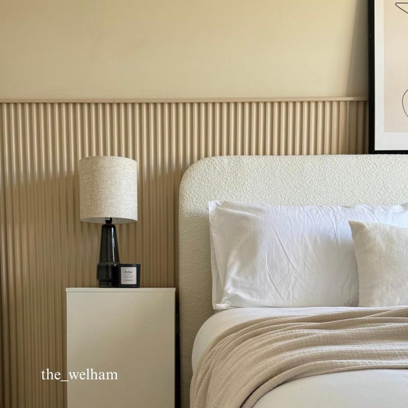 May include: A white bed with a fluffy headboard and a beige blanket. A black lamp with a beige lampshade sits on a white bedside table next to the bed. The wall behind the bed is beige and has a wood paneling design.