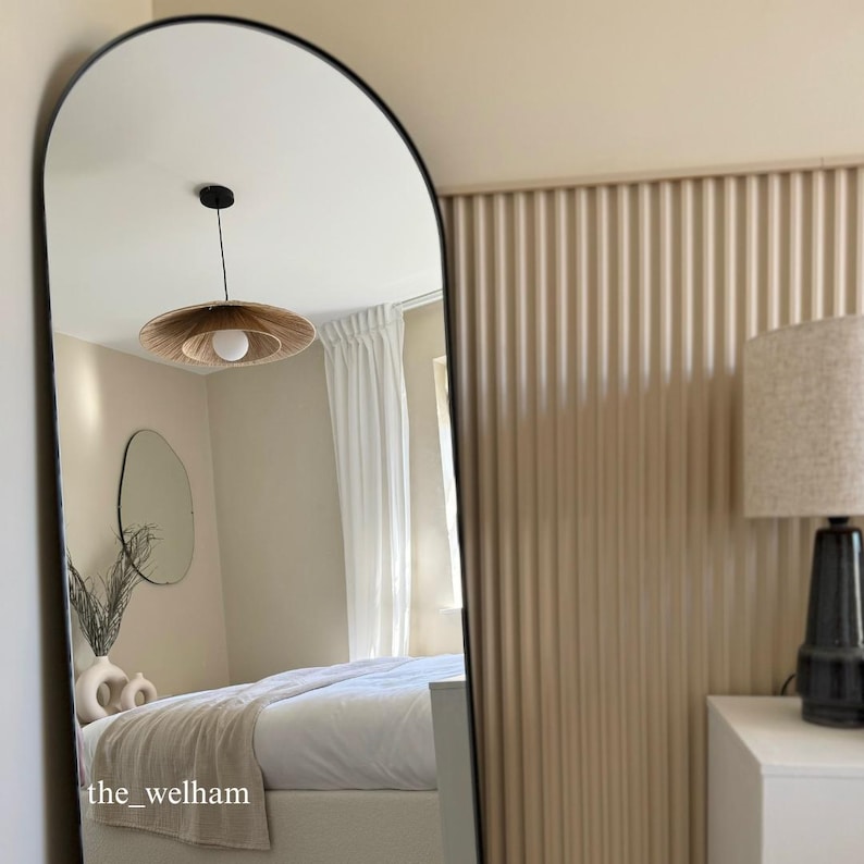 May include: A large arched mirror with a black frame reflects a bedroom with a white bed, a white ceiling, and a light fixture with a woven shade. The wall behind the mirror is a light beige colour with vertical grooves.