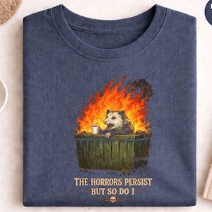 Könnte beinhalten: Marineblaues Heather-T-Shirt mit einem Grafik-Design eines Opossums in einem brennenden Müllcontainer, das eine Kaffeetasse hält. Der Text "THE HORRORS PERSIST BUT SO DO I" ist darunter. Rundhalsausschnitt.