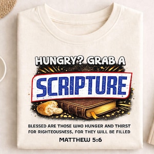 Op de afbeelding: Crèmekleurig T-shirt met de tekst "HUNGRY? GRAB A SCRIPTURE" in een kleurrijk ontwerp. Daaronder een afbeelding van een boek en brood. Het shirt bevat ook de tekst "BLESSED ARE THOSE WHO HUNGER AND THIRST FOR RIGHTEOUSNESS, FOR THEY WILL BE FILLED MATTHEW 5:6."