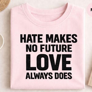 Może przedstawiać: Jasnoróżowa koszulka z napisem "HATE MAKES NO FUTURE LOVE ALWAYS DOES" czarnymi, pogrubionymi literami. Koszulka jest złożona na jasnej powierzchni, z notatnikiem i długopisem.
