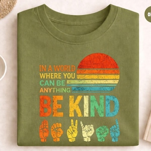 Pode incluir: T-shirt verde-oliva com o texto "IN A WORLD WHERE YOU CAN BE ANYTHING BE KIND" num design retro. O design inclui um pôr do sol colorido e símbolos de linguagem gestual.