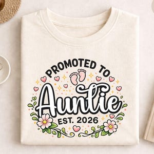 May include: A cream-coloured sweatshirt featuring the text "Promoted to Auntie EST. 2026". The design includes pink hearts, baby footprints, and floral accents. The word "NATURAL" is printed on the upper right corner of the sweatshirt.