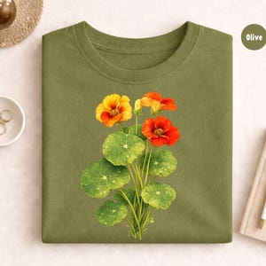 May include: Olive green t-shirt featuring a botanical illustration of orange and yellow nasturtium flowers with green leaves. The flowers have water droplets on the leaves. The word "Olive" is in a circle on the top right.