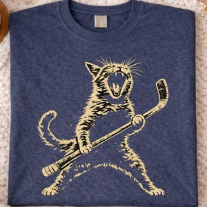 Könnte beinhalten: Marineblaues T-Shirt mit einer beigen Illustration einer Katze, die einen Hockeyschläger hält und brüllt. Das Shirt besteht aus einem weichen Material. Der Text "Heather Navy" befindet sich in der oberen rechten Ecke.