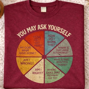 May include: Maroon t-shirt with the text "YOU MAY ASK YOURSELF" in a circular design. The design includes questions like "How did I get here?" and "Where is that large automobile?" on a soft, textured surface.