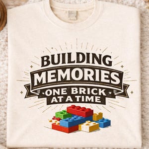 Op de afbeelding: Crèmekleurige sweatshirt met de tekst "BUILDING MEMORIES ONE BRICK AT A TIME" in zwart en goud. Het ontwerp bevat kleurrijke speelgoedbouwstenen. De sweatshirt wordt getoond op een gezellig, getextureerd oppervlak.
