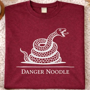 Op de afbeelding: Bordeauxrood t-shirt met een witte afbeelding van een opgerolde slang met uitgestoken tong. De tekst "DANGER NOODLE" staat eronder. Het shirt ligt op een witte, getextureerde ondergrond.