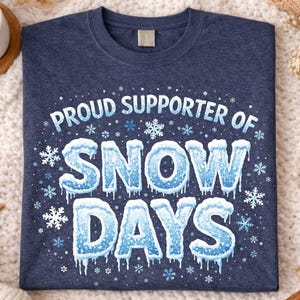 May include: Heather navy blue t-shirt with the text "Proud Supporter of Snow Days" in a wintery design. The text is in white and blue, with snowflakes and icicles. The shirt is displayed on a cozy, white, textured surface.
