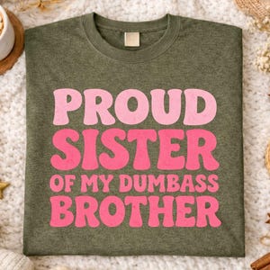 May include: Olive green t-shirt with the text "PROUD SISTER OF MY DUMBASS BROTHER" in pink bubble letters. The shirt is displayed on a white, textured surface with decorative elements.