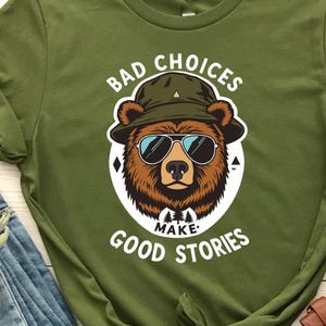 Könnte beinhalten: Olivgrünes T-Shirt mit einem Bären-Motiv, das eine Sonnenbrille und einen Hut trägt, mit dem Text "BAD CHOICES MAKE GOOD STORIES". Das Shirt wird mit blauen Jeans und braunen Stiefeletten kombiniert.