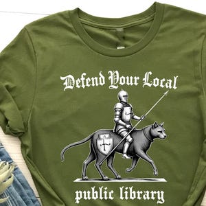 May include: Olive green t-shirt with the text "Defend Your Local Public Library" and a graphic of a knight riding a wolf-like creature. The shirt is paired with blue jeans and brown ankle boots.