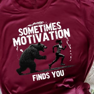 Könnte beinhalten: Burgunderfarbenes T-Shirt mit dem Text "Sometimes Motivation Finds You" und einer Grafik eines Bären, der einen Läufer verfolgt. Das Shirt hat eine einfarbige burgunderrote Farbe und einen Rundhalsausschnitt.