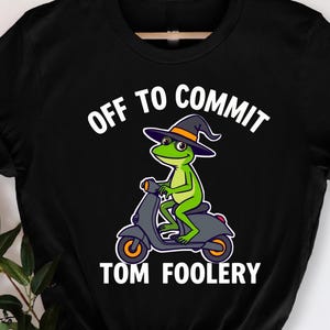 Op de afbeelding: Zwart t-shirt met een groene kikker met een heksenhoed op een scooter. De tekst "OFF TO COMMIT TOM FOOLERY" staat boven en onder de kikkerillustratie in het wit.