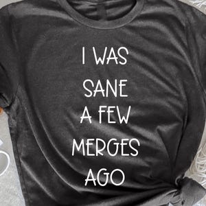 Könnte beinhalten: Dunkelgraues Melange-T-Shirt mit dem weißen Text "I WAS SANE A FEW MERGES AGO". Das Shirt wird auf einer weißen Oberfläche mit einer weißen Tasse, einer gewebten Matte und einem Zweig Blumen präsentiert.