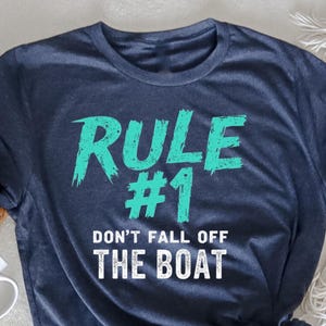 Puede incluir: Una camiseta azul marino jaspeada con el texto "RULE #1 DON'T FALL OFF THE BOAT" en letras blancas y turquesa.