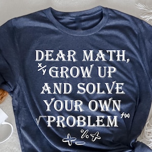 Könnte beinhalten: Ein marineblaues T-Shirt mit dem Text "Liebe Mathe, werde erwachsen und löse dein eigenes Problem" in Weiß. Der Text ist von mathematischen Symbolen umgeben.