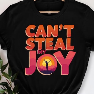 May include: Black t-shirt with the text "CAN'T STEAL MY JOY" in orange and pink lettering. The design includes a silhouette of a person with arms raised in front of a sunset. The shirt is hanging on a wooden hanger.