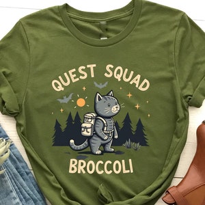 Könnte beinhalten: Olivengrünes T-Shirt mit einer Comic-Katze, die einen Rucksack trägt, und dem Text "Quest Squad Broccoli".