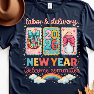Op de afbeelding: Marineblauw t-shirt met een kleurrijk grafisch ontwerp. Het ontwerp bevat de tekst "labor & delivery", "2026", "NEW YEAR" en "welcome committee". De afbeelding toont ook een wereldbol, een babyfles en een regenboog.