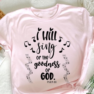 K&ouml;nnte beinhalten: Hellrosa T-Shirt mit dem schwarzen Schriftzug "I will sing of the goodness of God", begleitet von Noten. Das Shirt zeigt auch den Bibelvers Psalm 89,1.